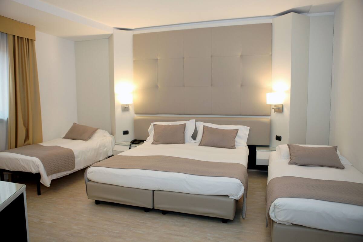 Habitación de hotel con dos camas individuales y una cama doble, todas con ropa de cama color beige. Hay dos mesitas de noche con lámparas, y un cabecero grande con panel de luz.