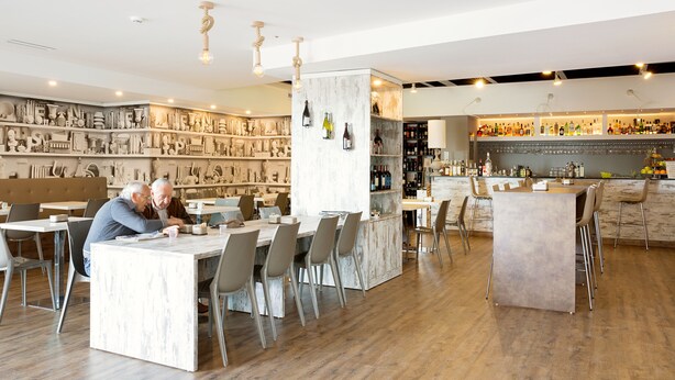 Un restaurante moderno con mesas y sillas de encimera de mármol, una zona de bar con una variedad de botellas y una pared decorativa con elementos escultóricos.