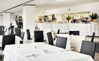 Un amplio comedor con varias mesas y sillas, un bufé central con diferentes opciones de comida, y una pared decorativa con arreglos florales.
