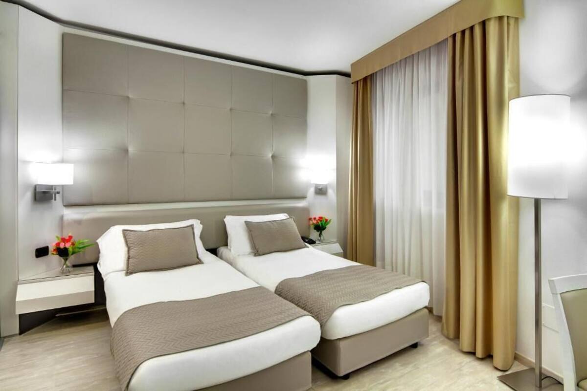 A hotel room with two beds, a large padded headboard, bedside lamps, a nightstand with a vase of flowers, and a desk with a lamp.
