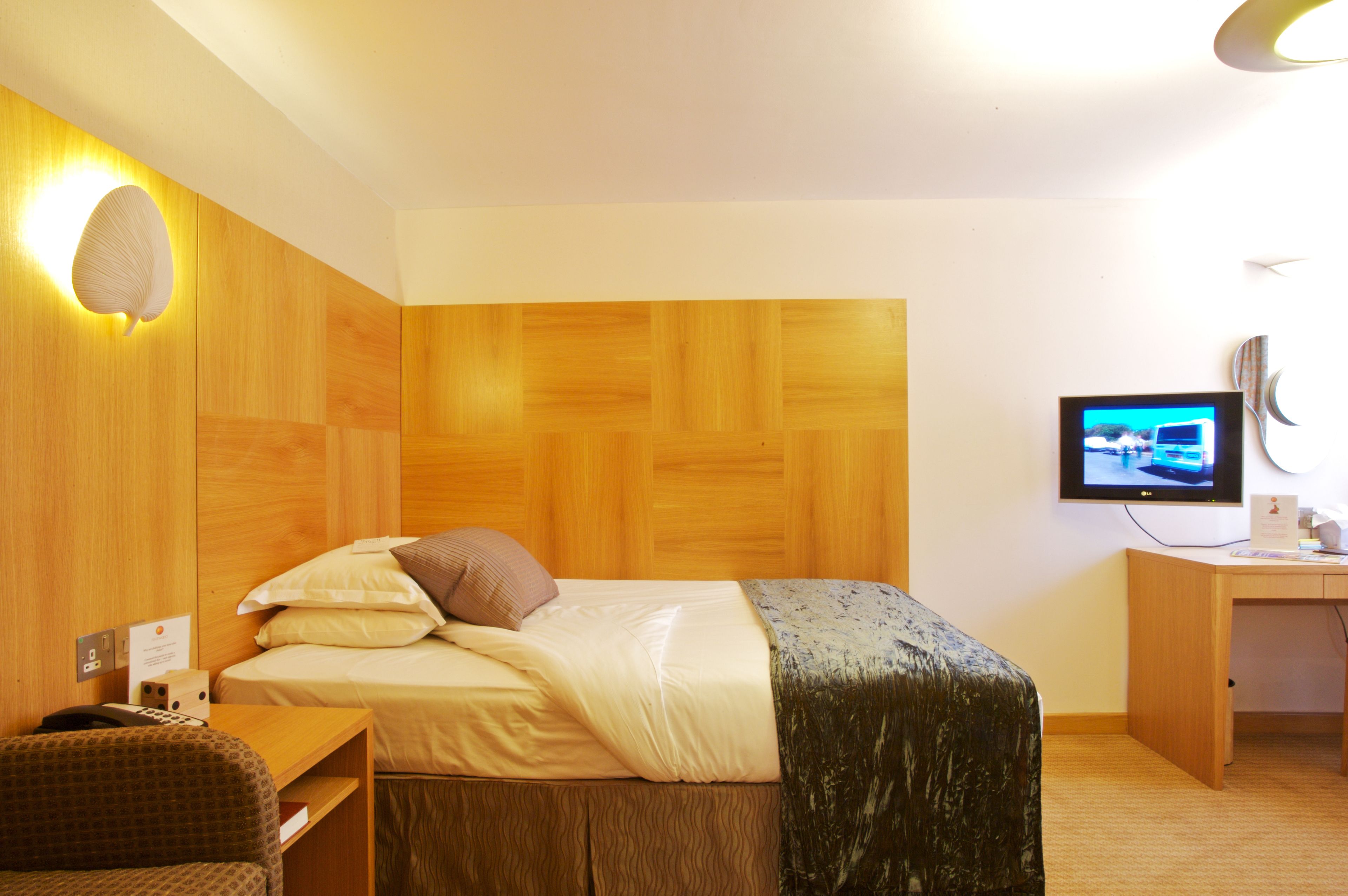 A hotel room with a wooden headboard, a bed with white linens, a desk with a computer, and a television mounted on the wall.
