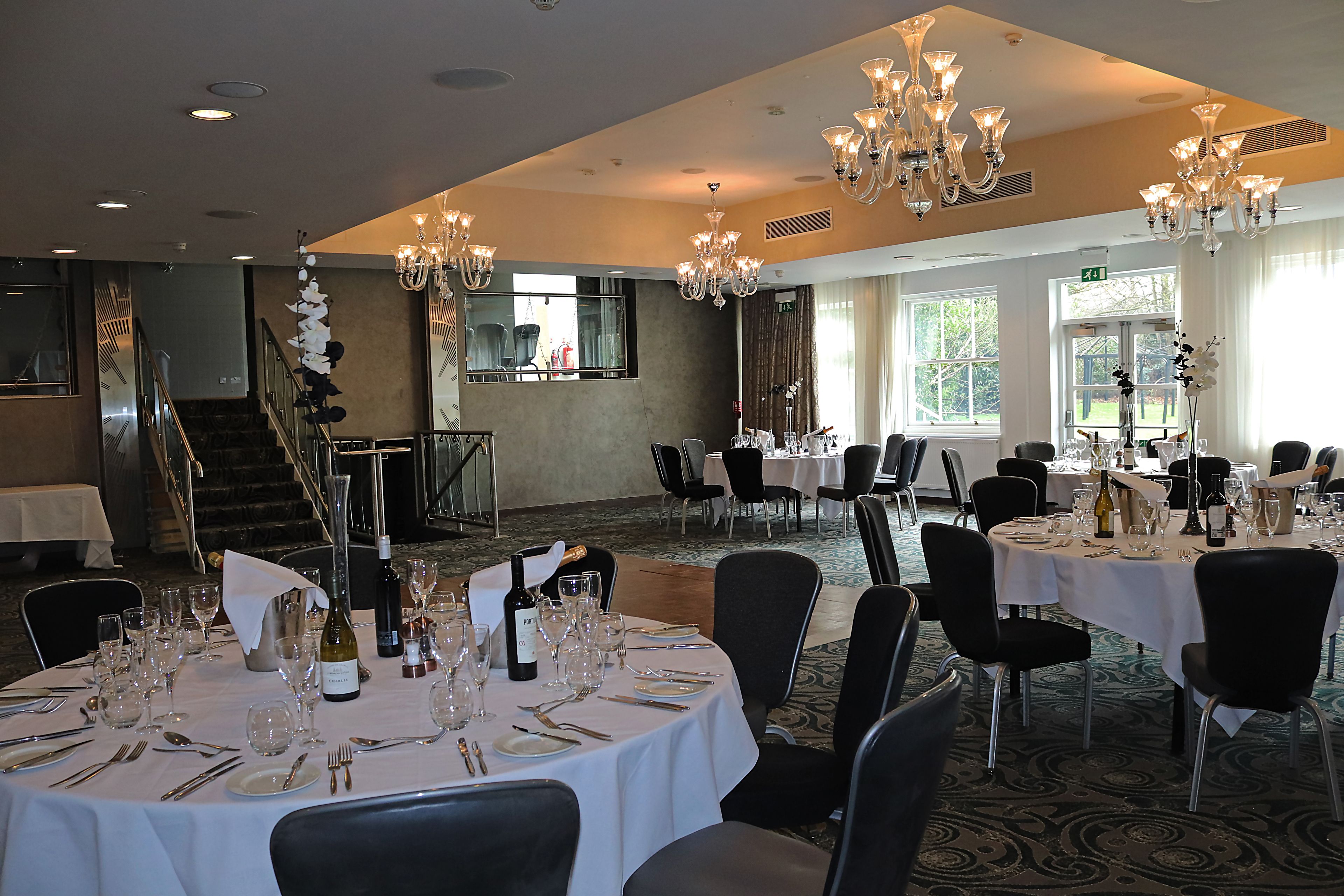A banquet hall with round tables set for a formal event, featuring chandeliers and a staircase in the background.