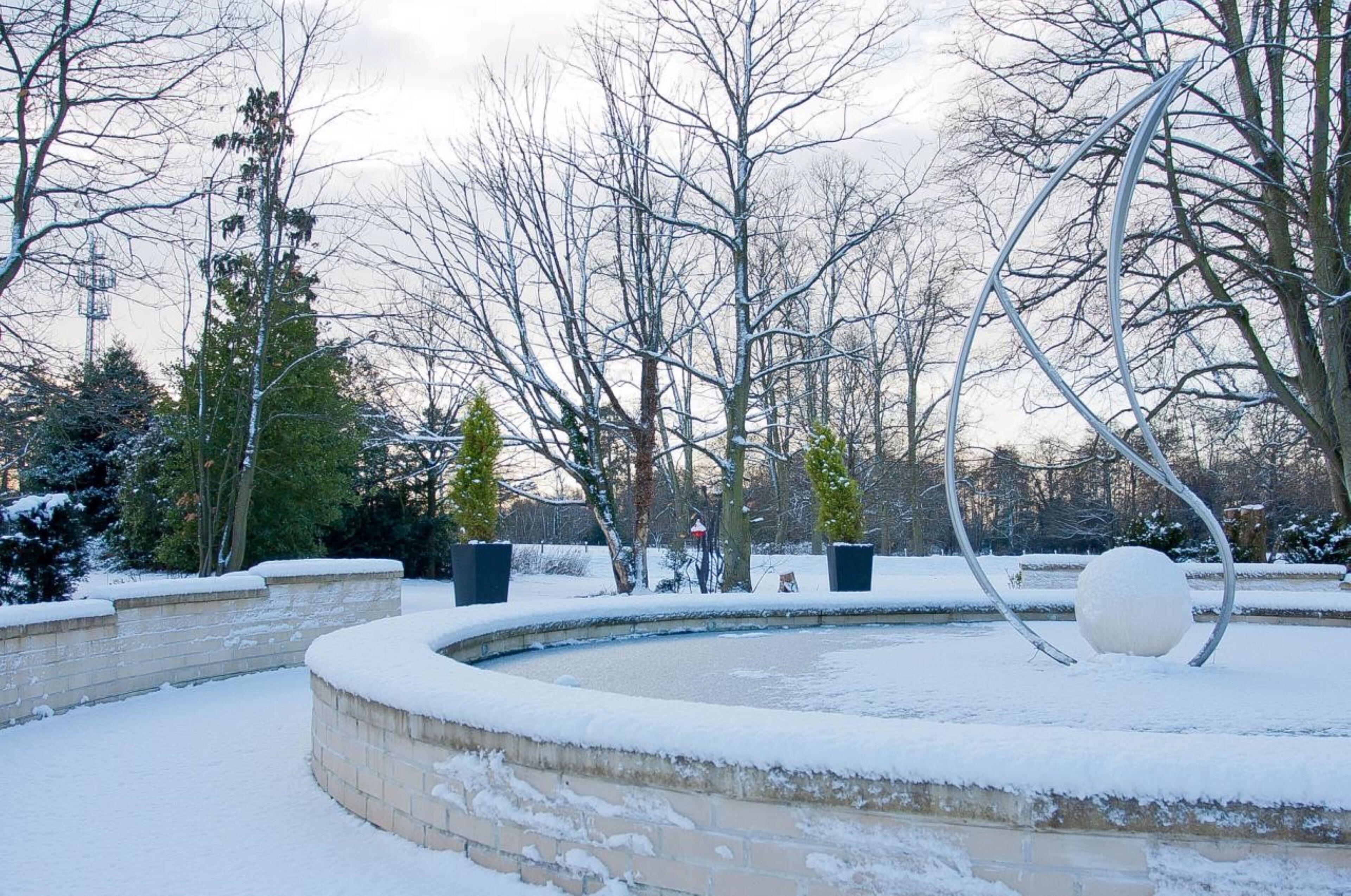 Een besneeuwde buitenruimte met een rond zwembad, metalen sculpturen en kale bomen.