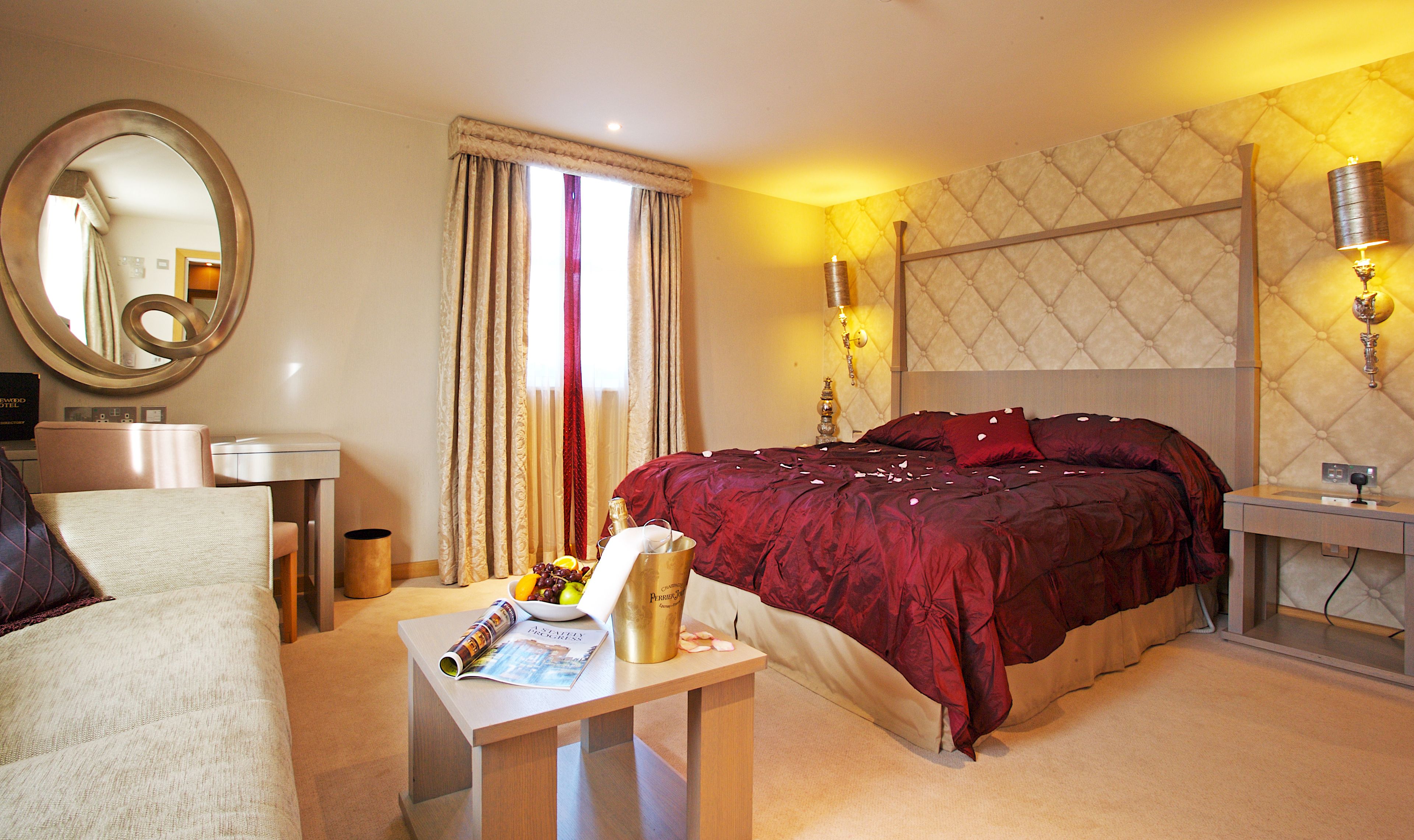 A hotel room with a bed, a desk with a lamp, a chair, a bedside table with a magazine, and a large mirror.