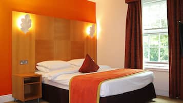 Habitación de hotel con cabecera de madera, cama con sábanas blancas y almohada roja, mesita de noche y ventana con cortinas.