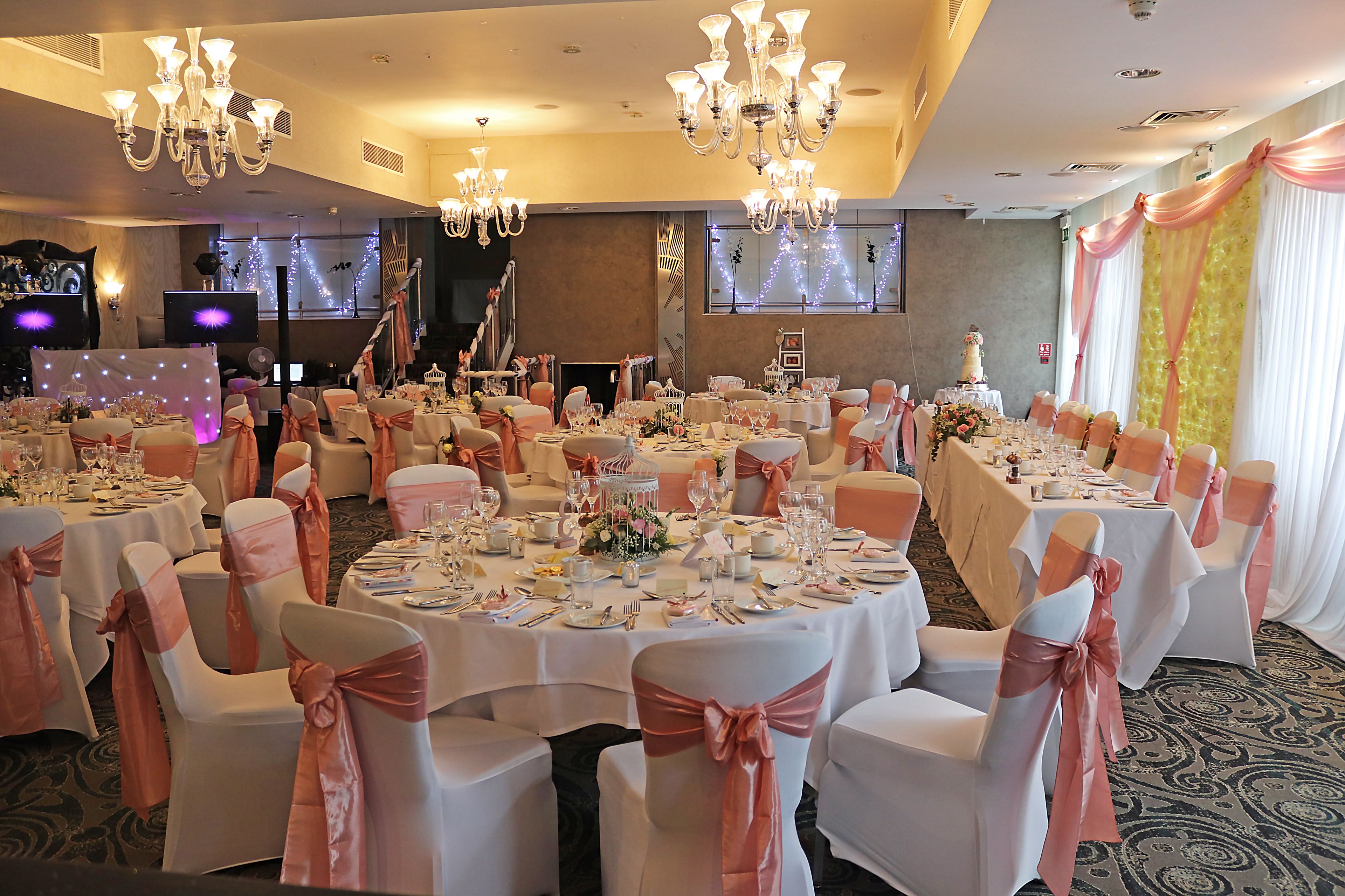 A banquet hall with round tables set for a formal event, adorned with white tablecloths and pink sashes.