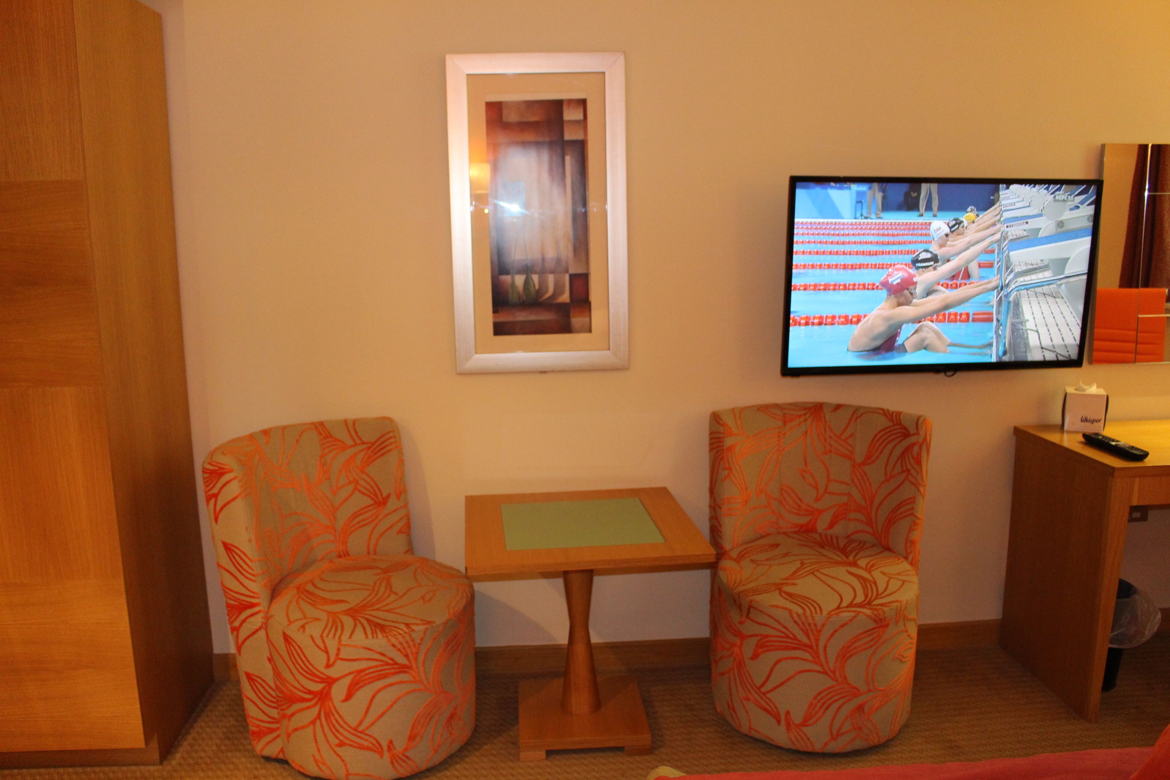 A hotel room with two patterned armchairs, a small table, a television displaying a swimming event, and a framed picture on the wall.