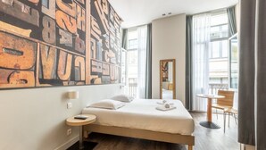 A hotel room with a large bed, a small round table, a chair, and a large abstract painting on the wall. - Urban Style Bordeaux Centre Hotel de la Presse (Bordeaux)