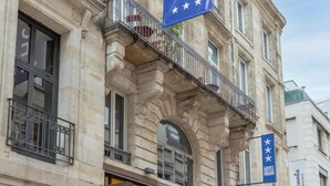 A hotel with a prominent blue flag and a storefront labeled "L'OCCITANE IN PROVENCE". - Urban Style Bordeaux Centre Hotel de la Presse (Bordeaux)