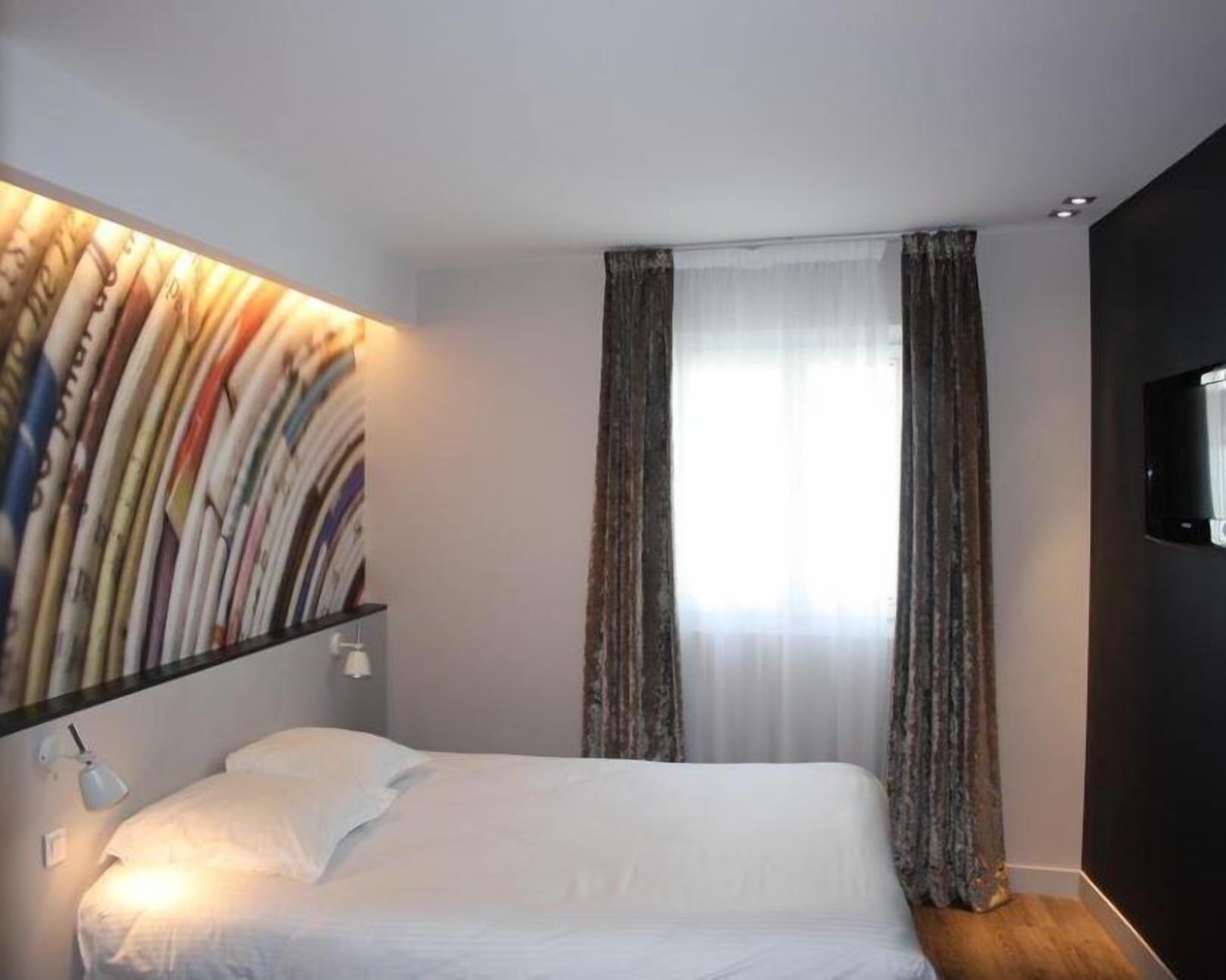 A hotel room with a bed, a wall-mounted shelf with books, a window with curtains, and a TV on the wall.