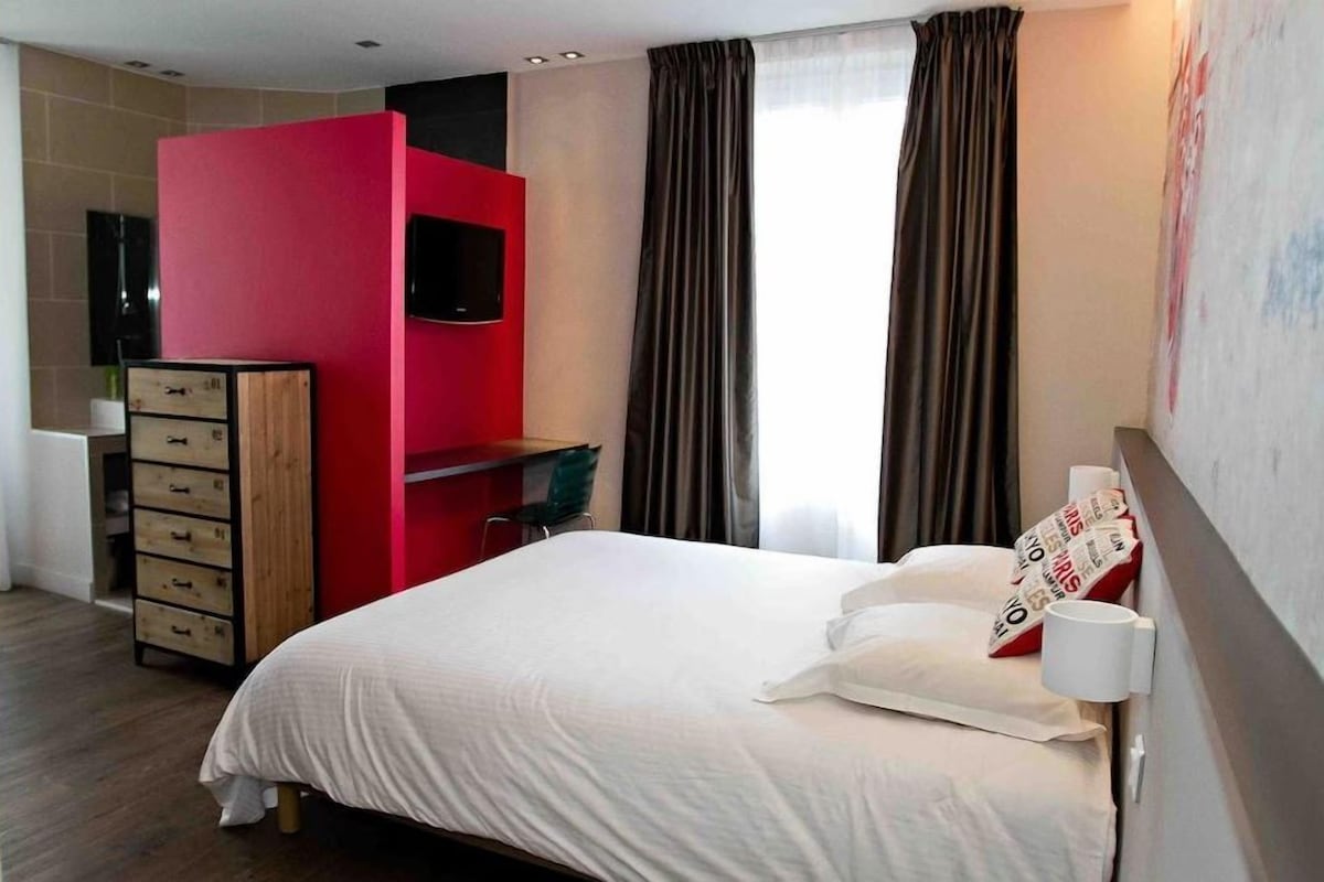 A hotel room with a bed, a red accent wall, a TV, a desk, and a window with curtains.