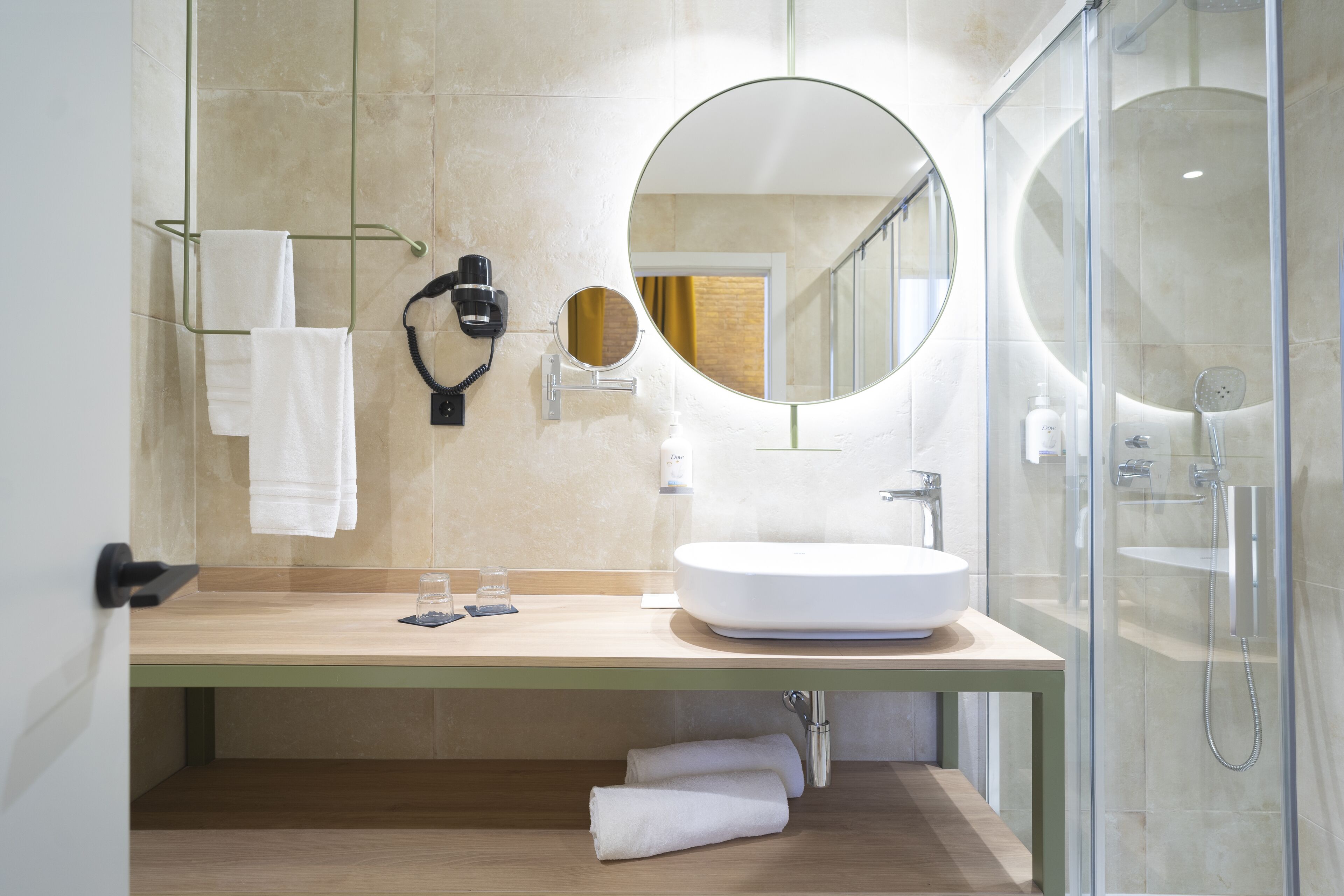 A modern bathroom with a large round mirror, a white sink, and a glass-enclosed shower.