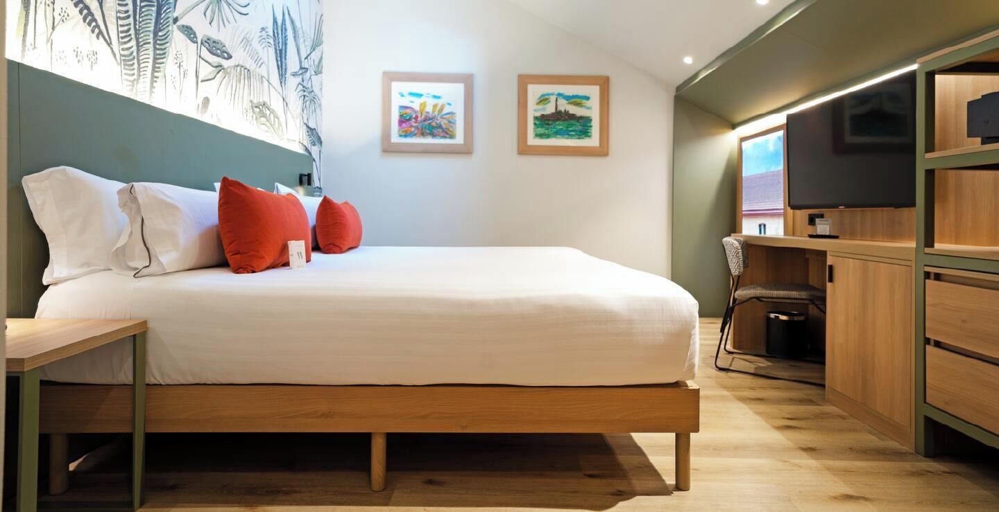 Un dormitorio con cama, almohadas, escritorio, silla y una obra de arte en la pared.