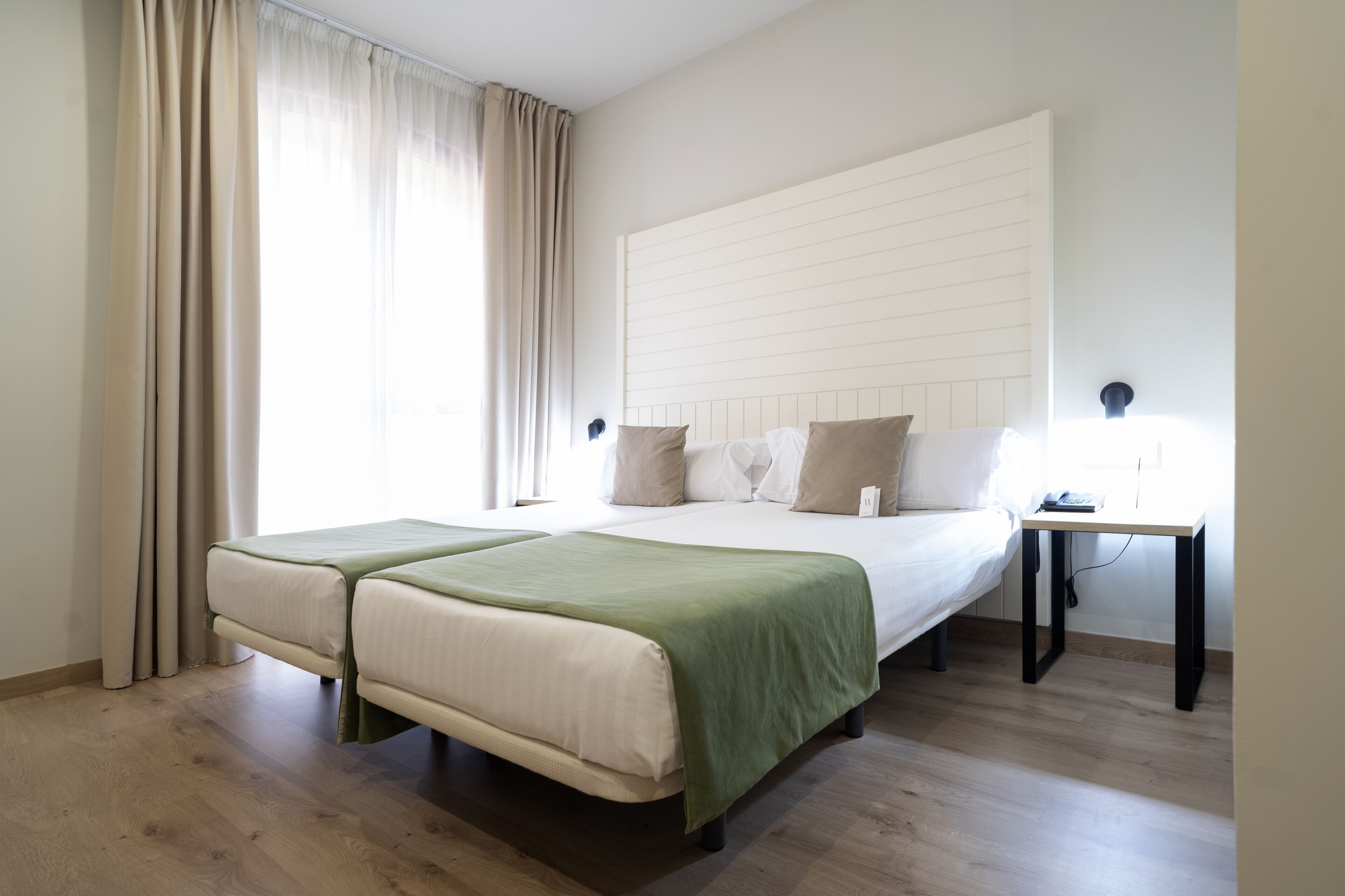 A double bed with a green bedspread and two pillows, a bedside table with a lamp, and a window with curtains.