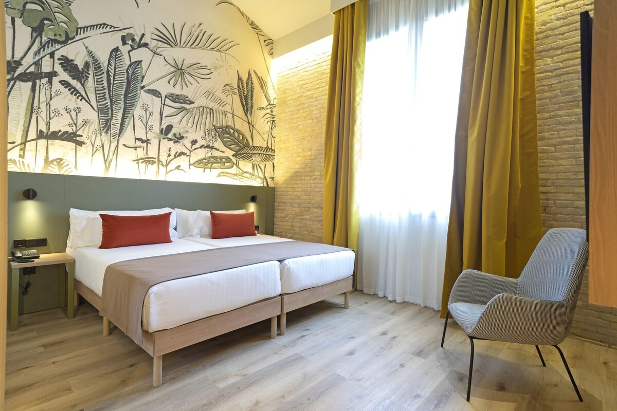A hotel room with a bed, a chair, a nightstand, and a wall mural.