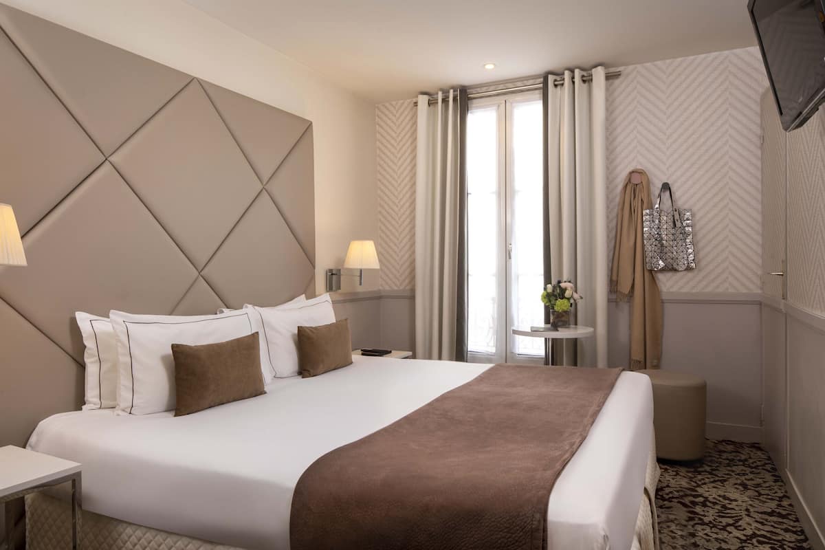 A hotel room with a large bed, a headboard with a geometric pattern, two bedside tables with lamps, a small round table with a vase of flowers, and a wardrobe with a mirror.