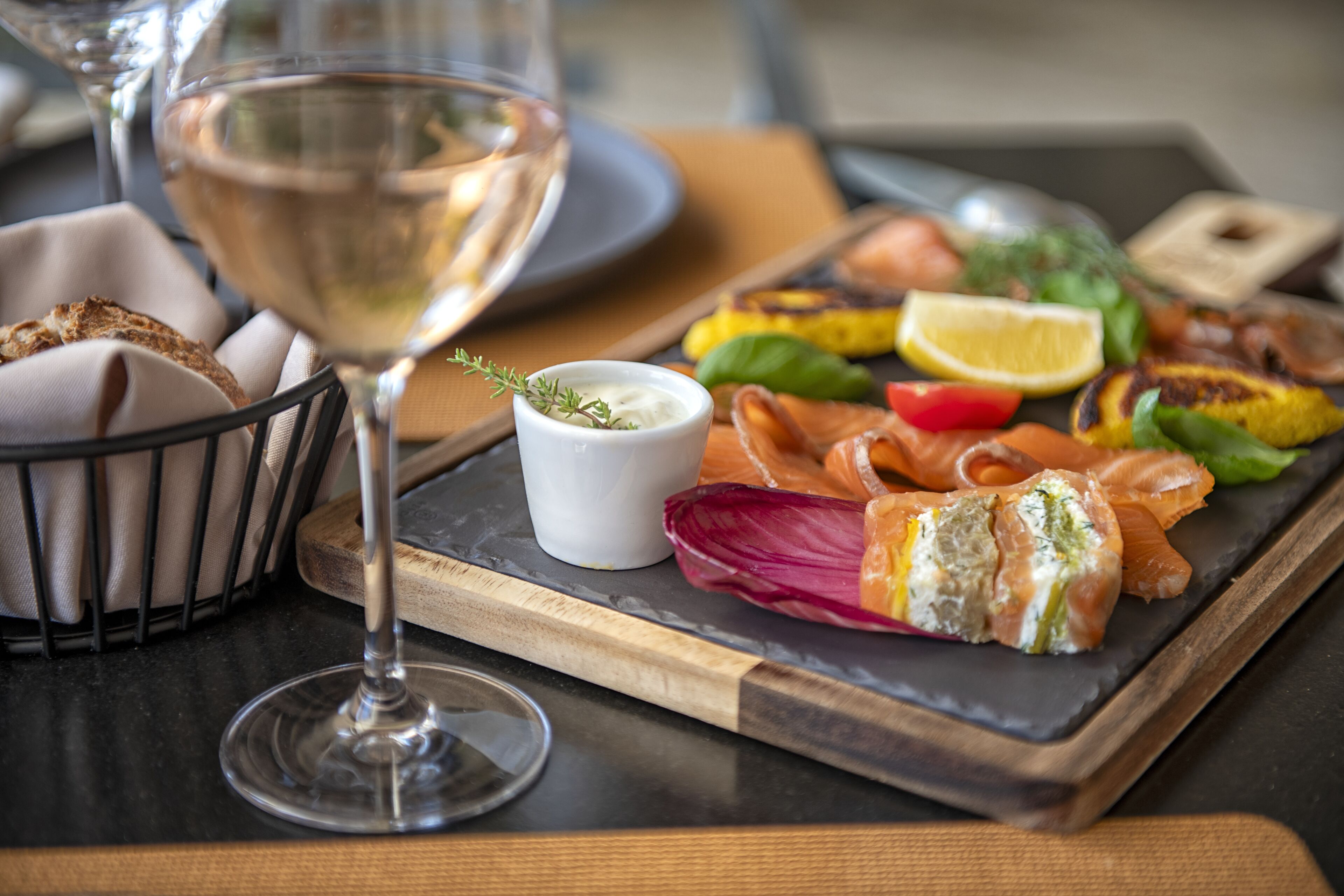 A dining table with a glass of white wine, a wooden board with various foods, and a small bowl of sauce.