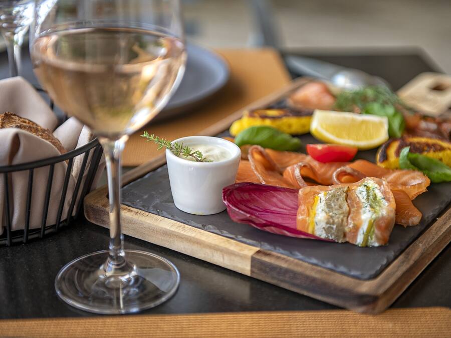 A dining table with a glass of white wine, a wooden board with various foods, and a small bowl of sauce.
