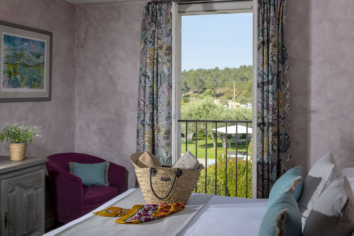 Une pièce comprenant un lit, un fauteuil violet, un panier en osier et une vue sur un paysage verdoyant par la fenêtre.
