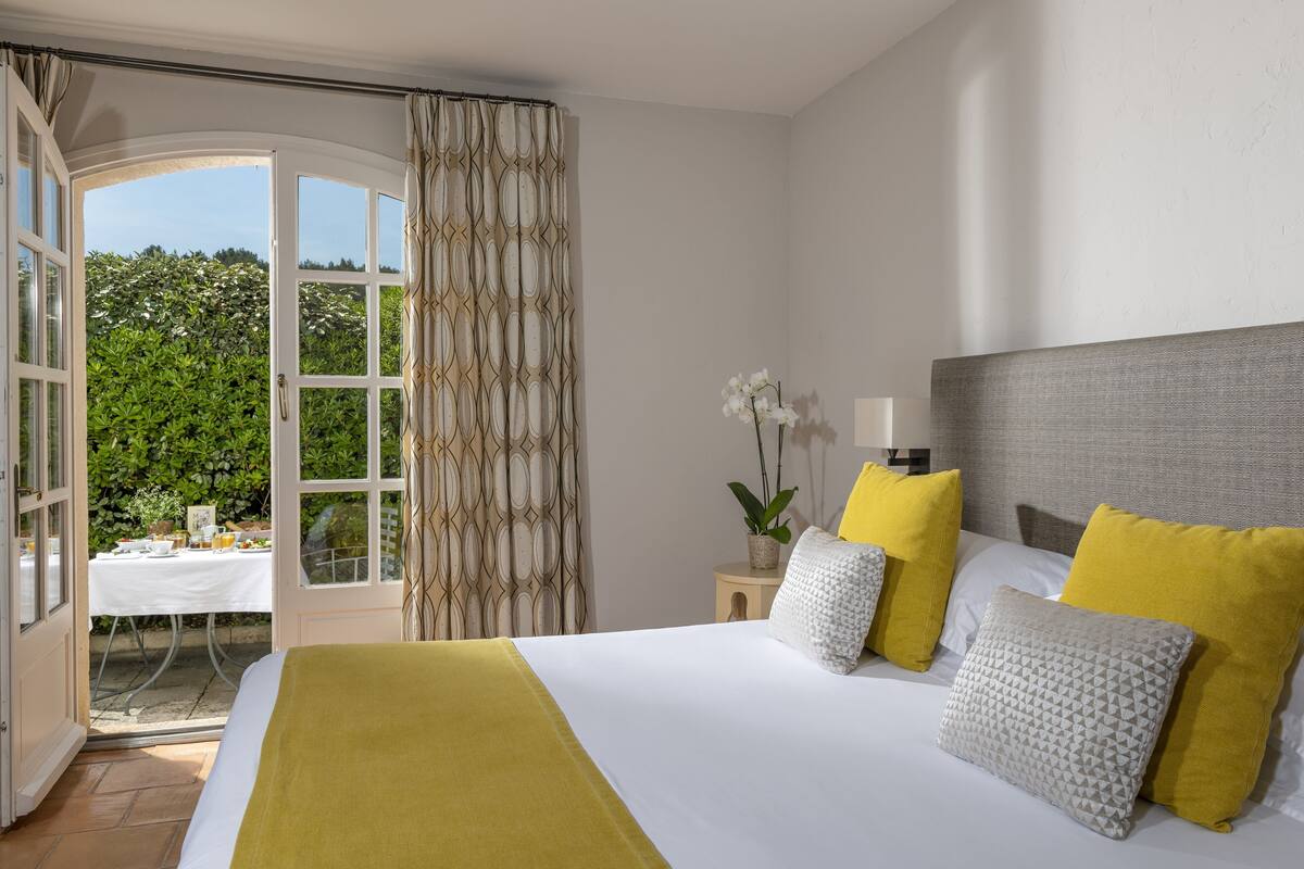 Une chambre à coucher avec un lit, des oreillers, une table de chevet, une lampe et une vue sur la verdure par une porte ouverte.