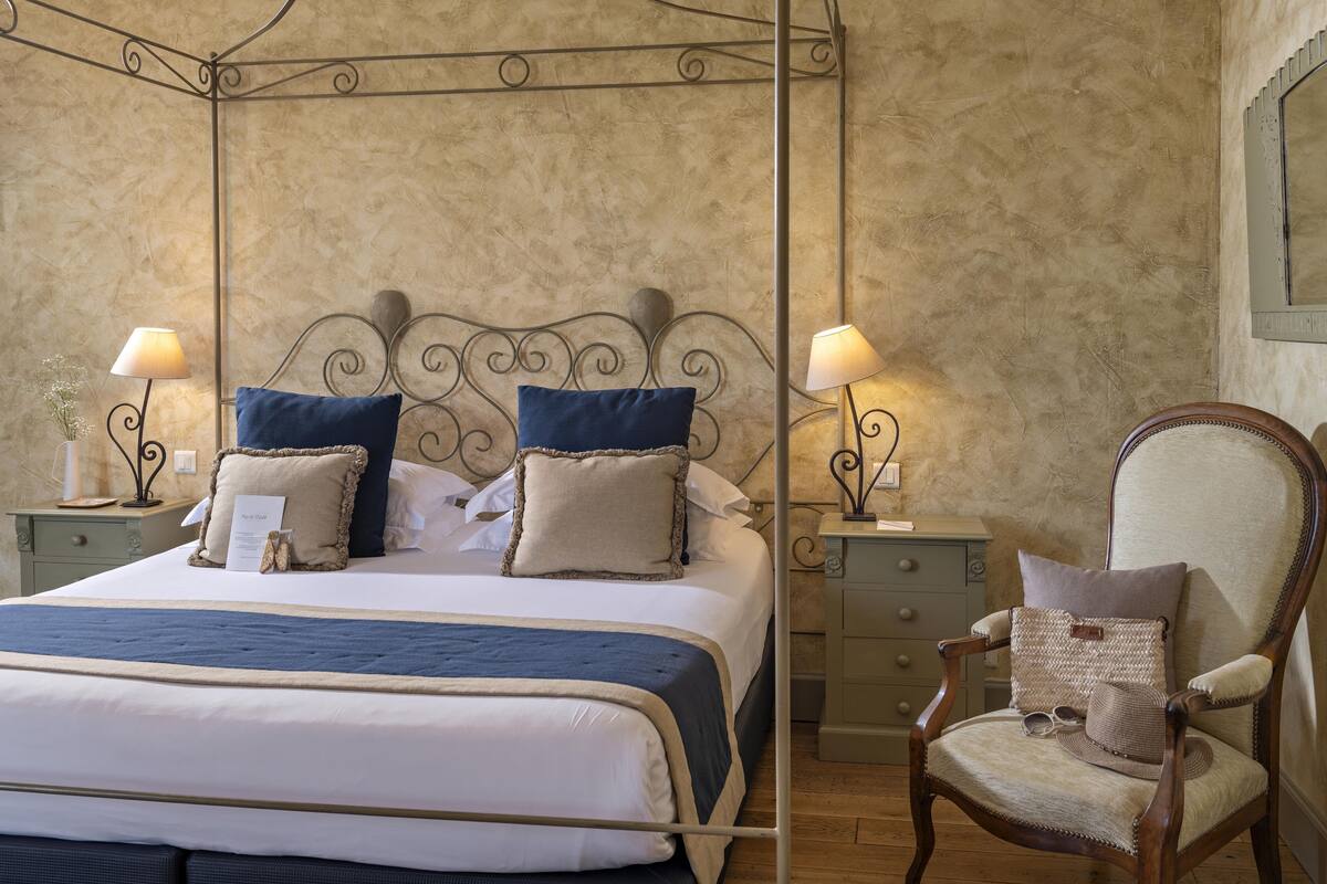 Un lit bien fait, avec une tête de lit, deux tables de chevet avec des lampes, une chaise avec un coussin et un miroir fixé au mur.