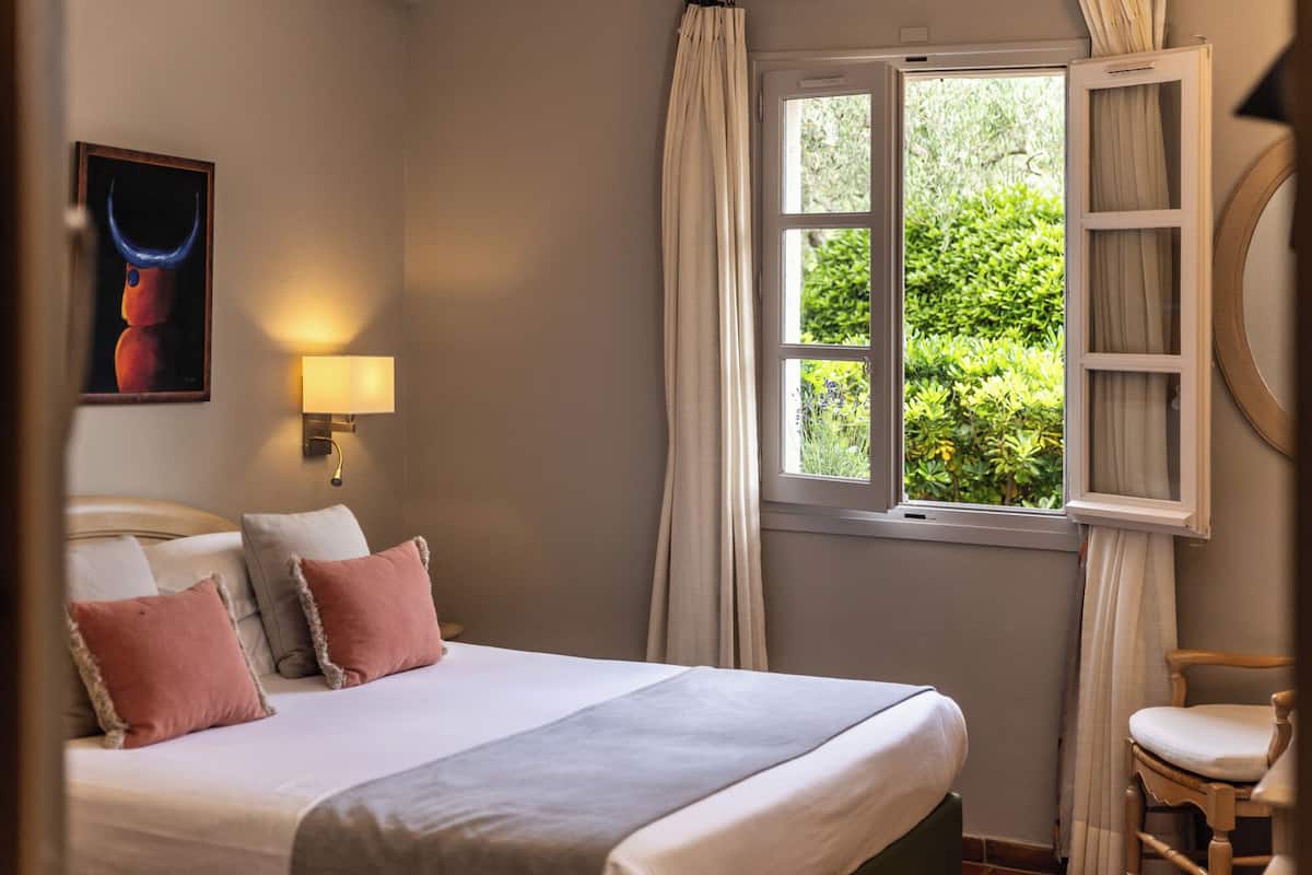 Une chambre à coucher avec un lit, une table de chevet, une lampe, une fenêtre avec des rideaux et un tableau au mur.