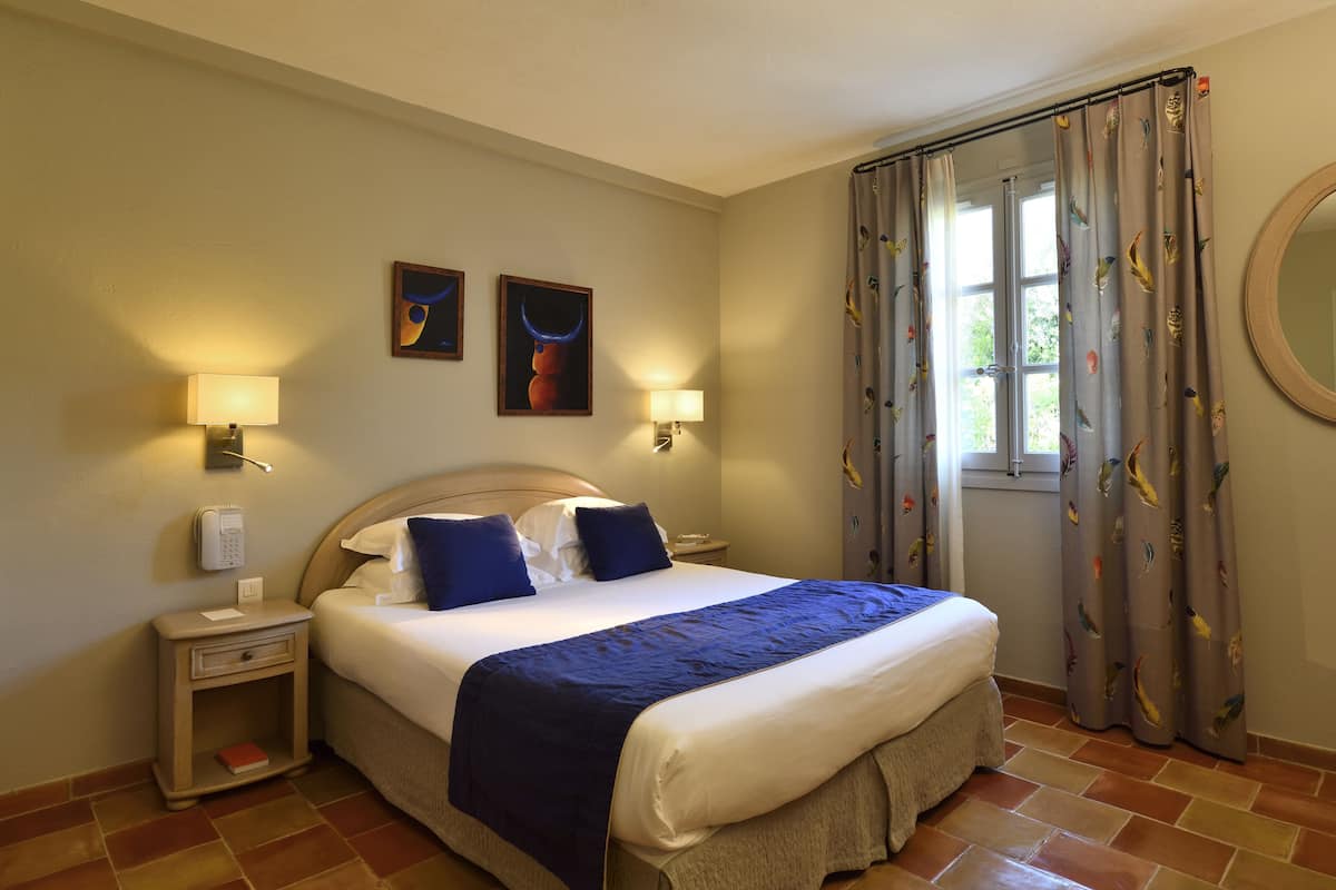 Une chambre d’hôtel comprenant un lit, deux lampes fixées au mur, une table de chevet, une commode, un miroir et une fenêtre avec des rideaux.