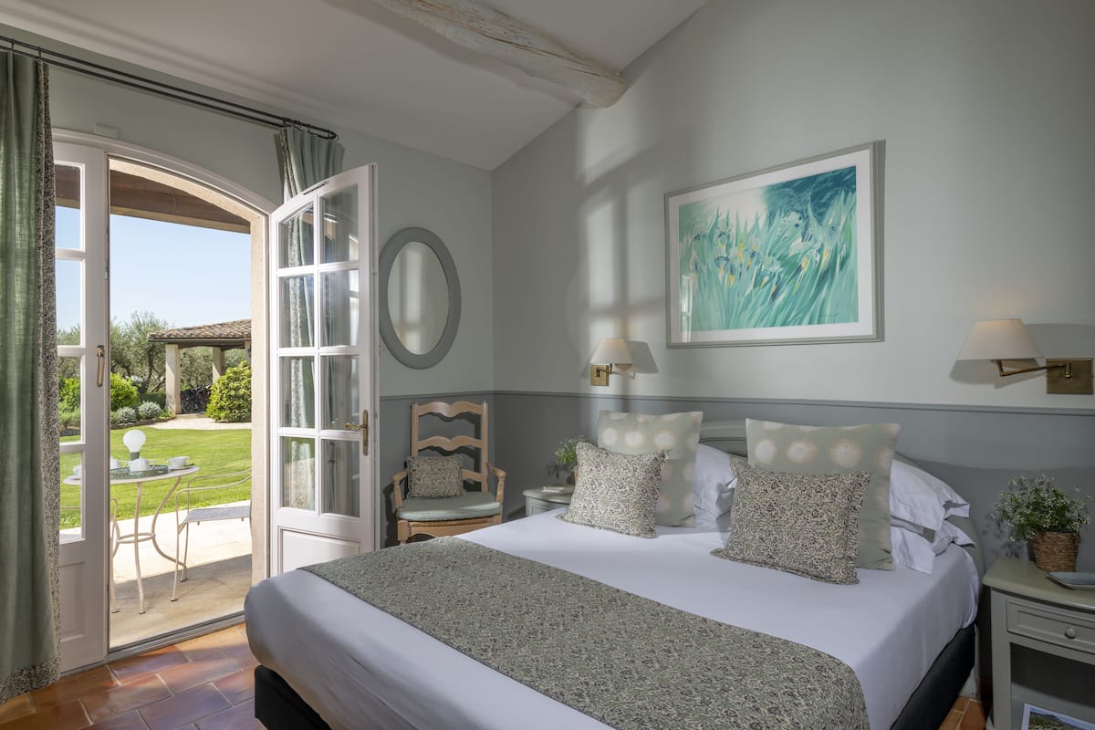 Une chambre avec un grand lit, une chaise, une table et une vue sur le jardin, accessible par une porte ouverte.