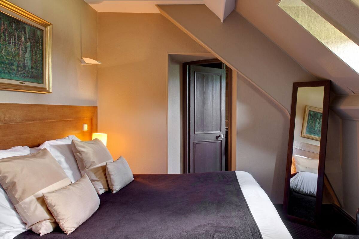 Et hotelværelse med en stor seng, et natbord med lampe, en dør og et spejl der afspejler en del af værelset.