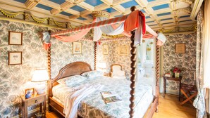 A four-poster bed with a canopy, a wooden nightstand with a lamp, and a floral wallpaper pattern. - Le Petit Manoir Des Bruyeres (Villefargeau)