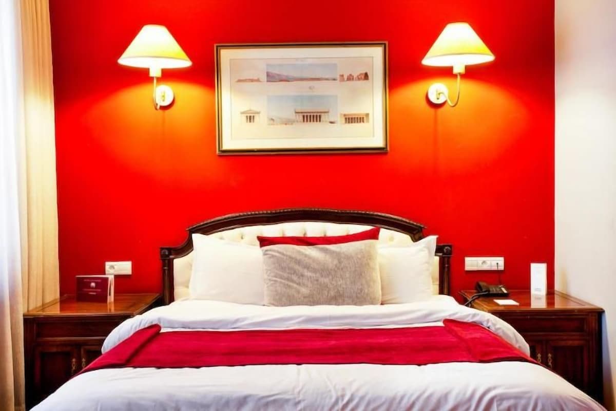 Een hotelkamer met een rode wand, een bed met wit beddengoed, twee bedlampen en een lijst met een afbeelding aan de wand.