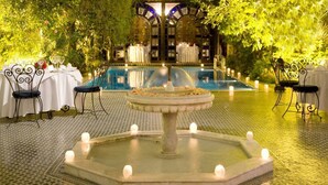 Zona junto a la piscina con una fuente, mesas y sillas, rodeada de vegetación.