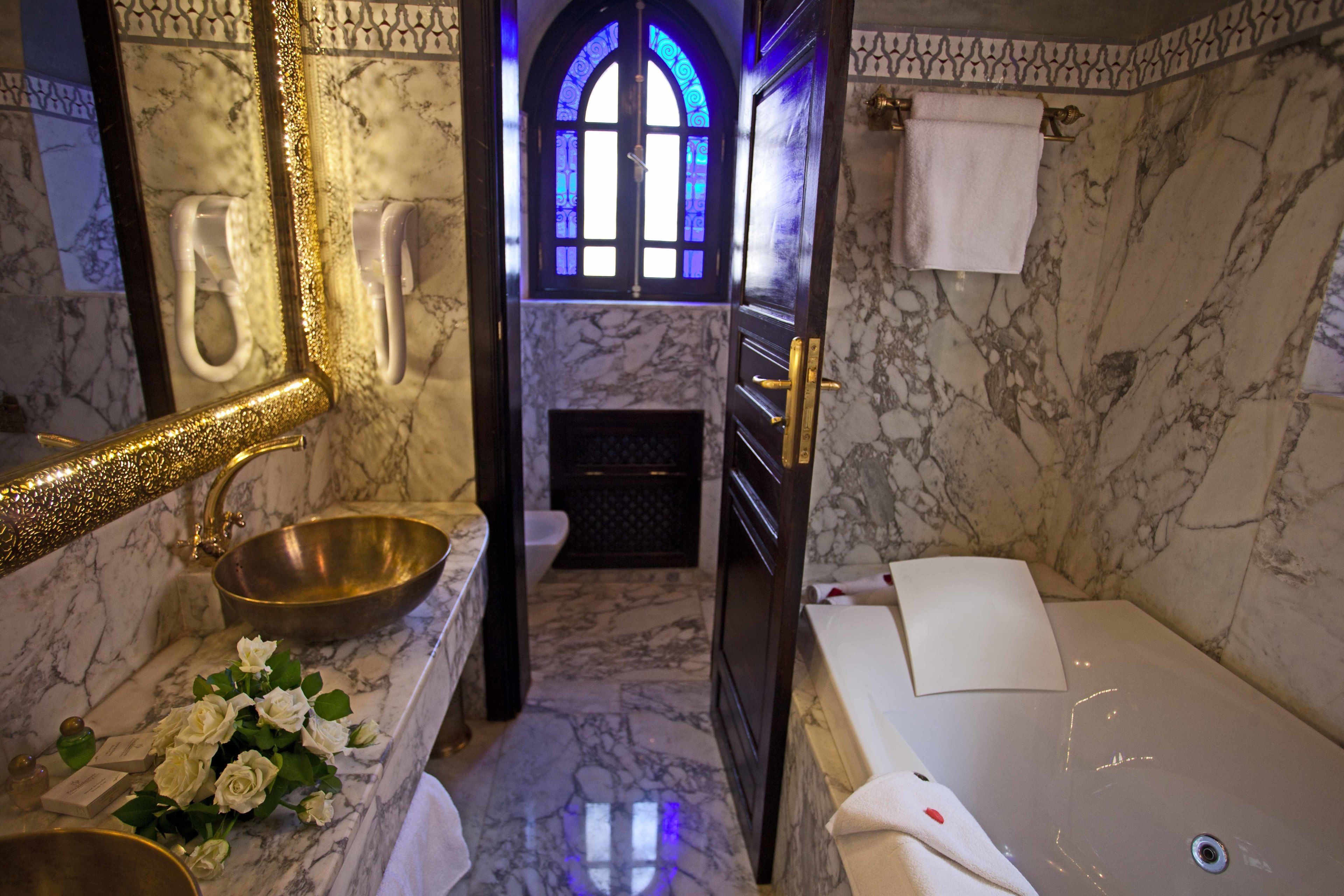 Une salle de bain avec un lavabo en marbre, un grand miroir, une baignoire et un vitrail.