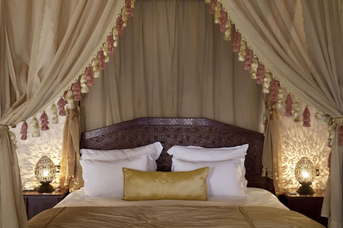 Una cama con almohadillas blancas y doradas, cabecera de madera y dosel con borlas colgantes.
