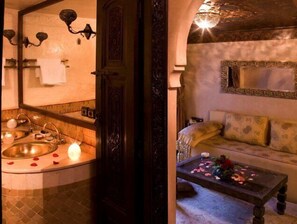 A bathroom with a round sink, two mirrors, and a chandelier. There is a sofa and a table with flowers in the adjacent room.