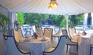 A tented outdoor dining area with tables set for a meal, surrounded by lush greenery and a view of a water feature.
