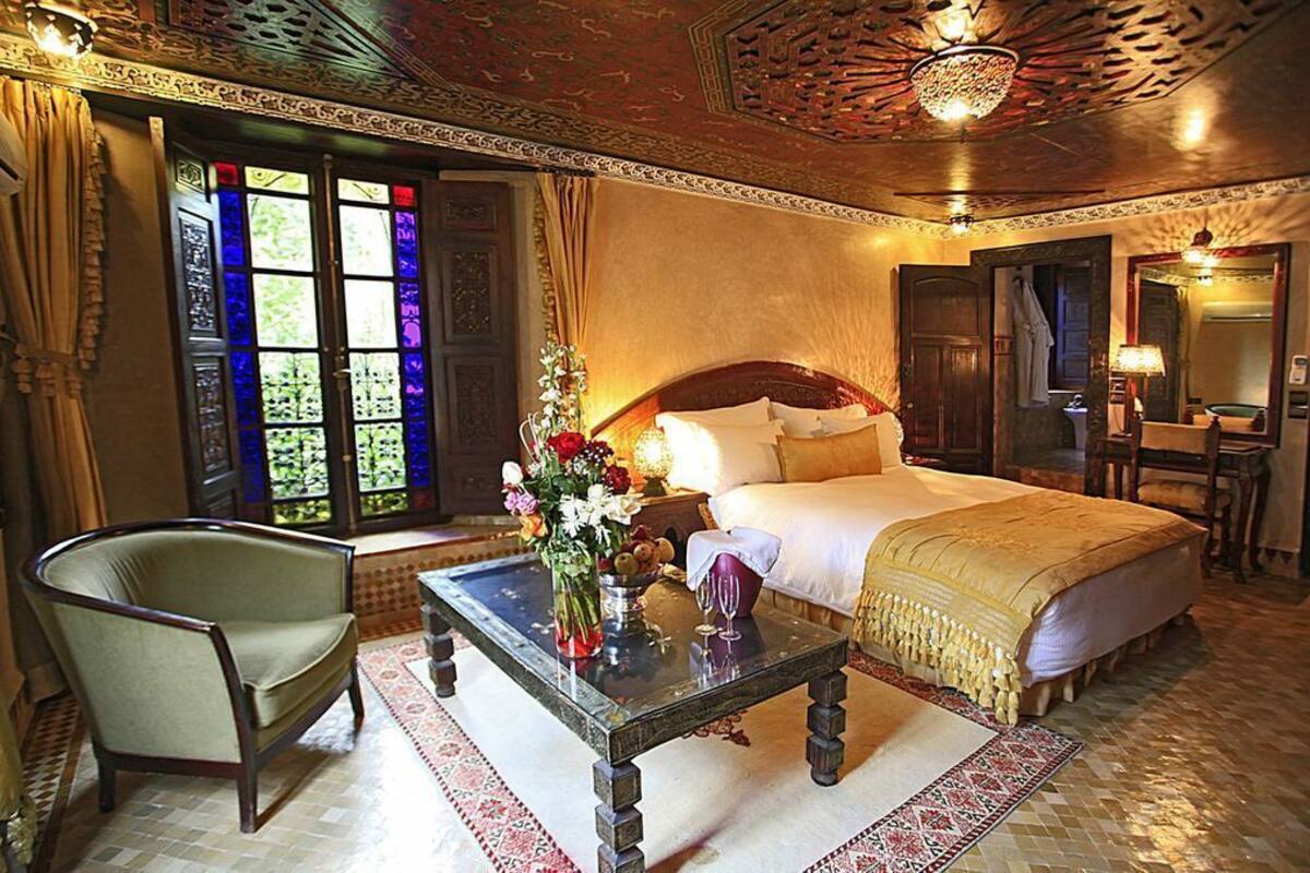 Una habitación tradicional con una cama grande, una silla de madera, una mesa de centro de vidrio con un jarrón de flores y vistas a un jardín a través de un ventanal de vitrales.