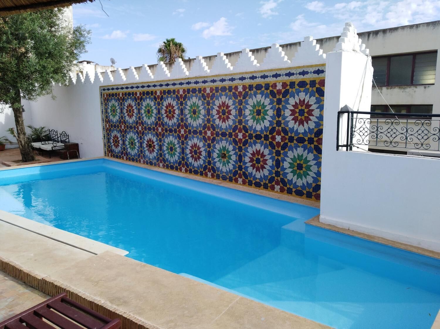 A swimming pool with a colorful mosaic wall, a white building, and a tree.