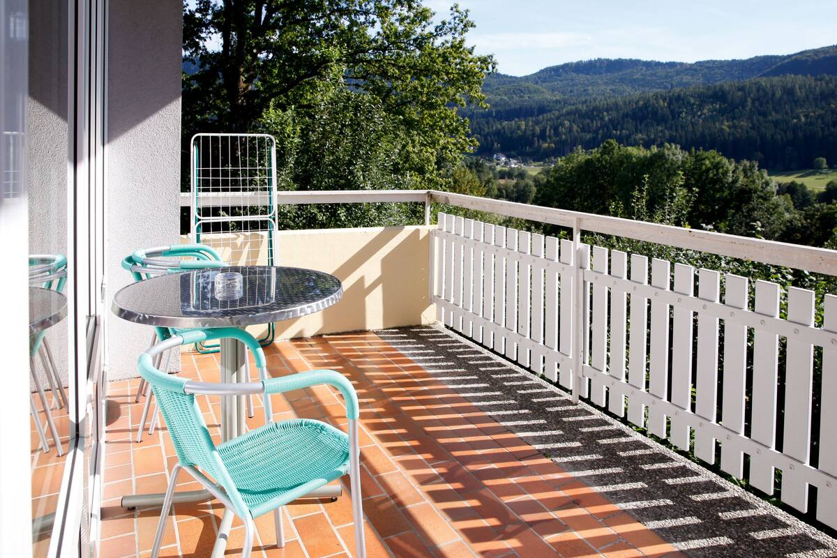 Un balcon avec une table et des chaises, une clôture blanche et une vue sur une vallée boisée.