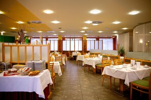 Een ruime eetruimte met meerdere tafels en stoelen, een buffetopstelling aan de linkerkant, en grote ramen voor natuurlijk licht.