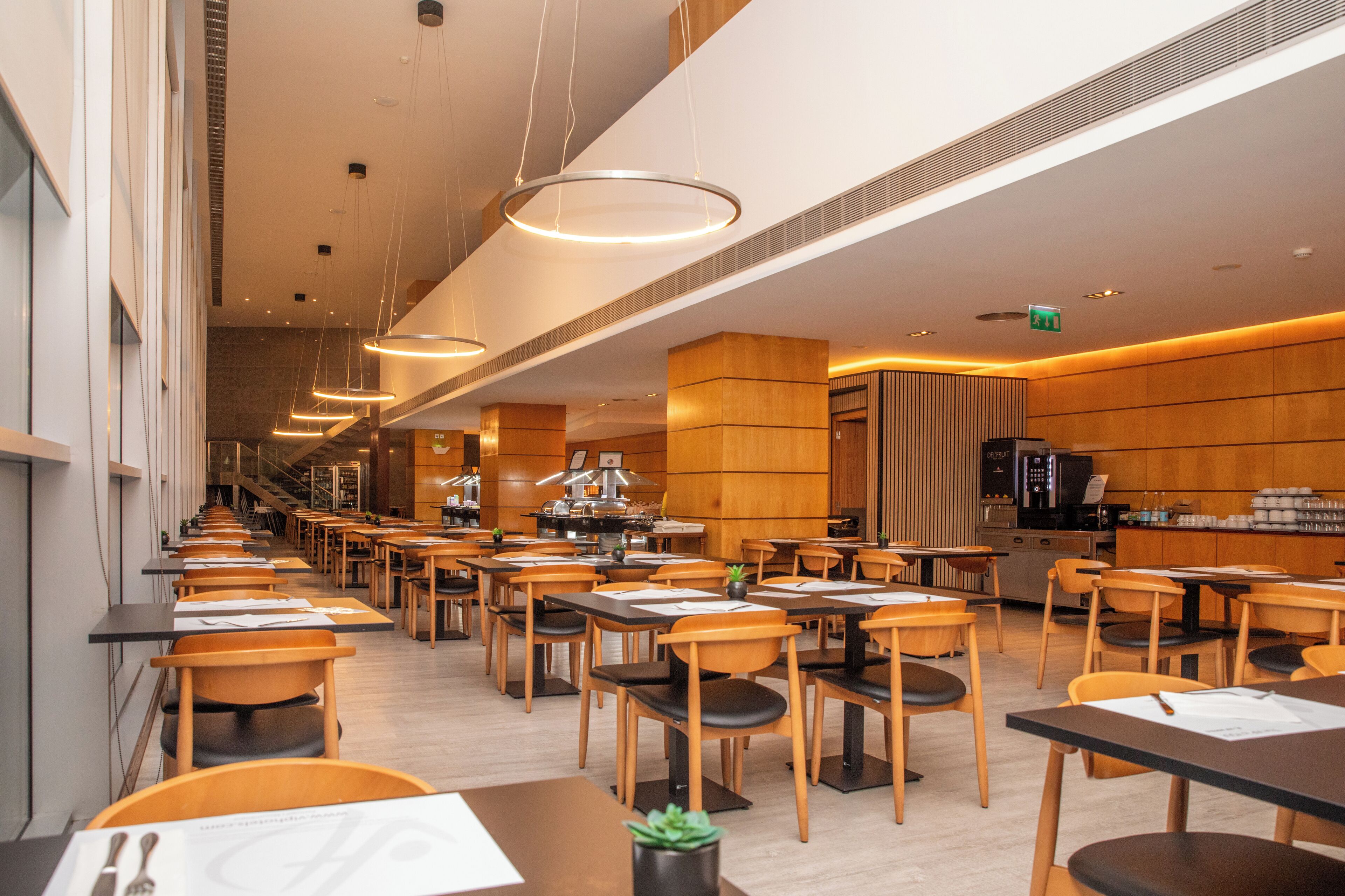 A modern restaurant with long tables and chairs, a kitchen area, and a clean, well-lit interior.