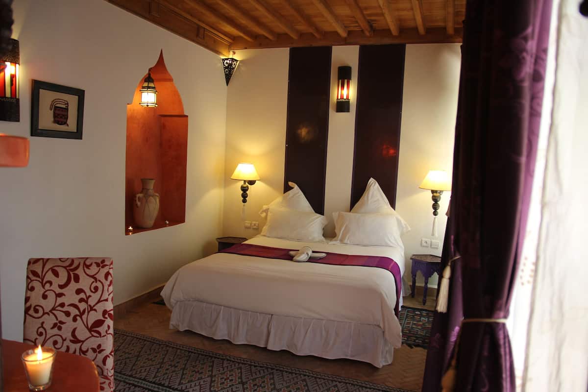A hotel room with a large bed, two bedside lamps, a nightstand, a decorative mirror, and a patterned chair.