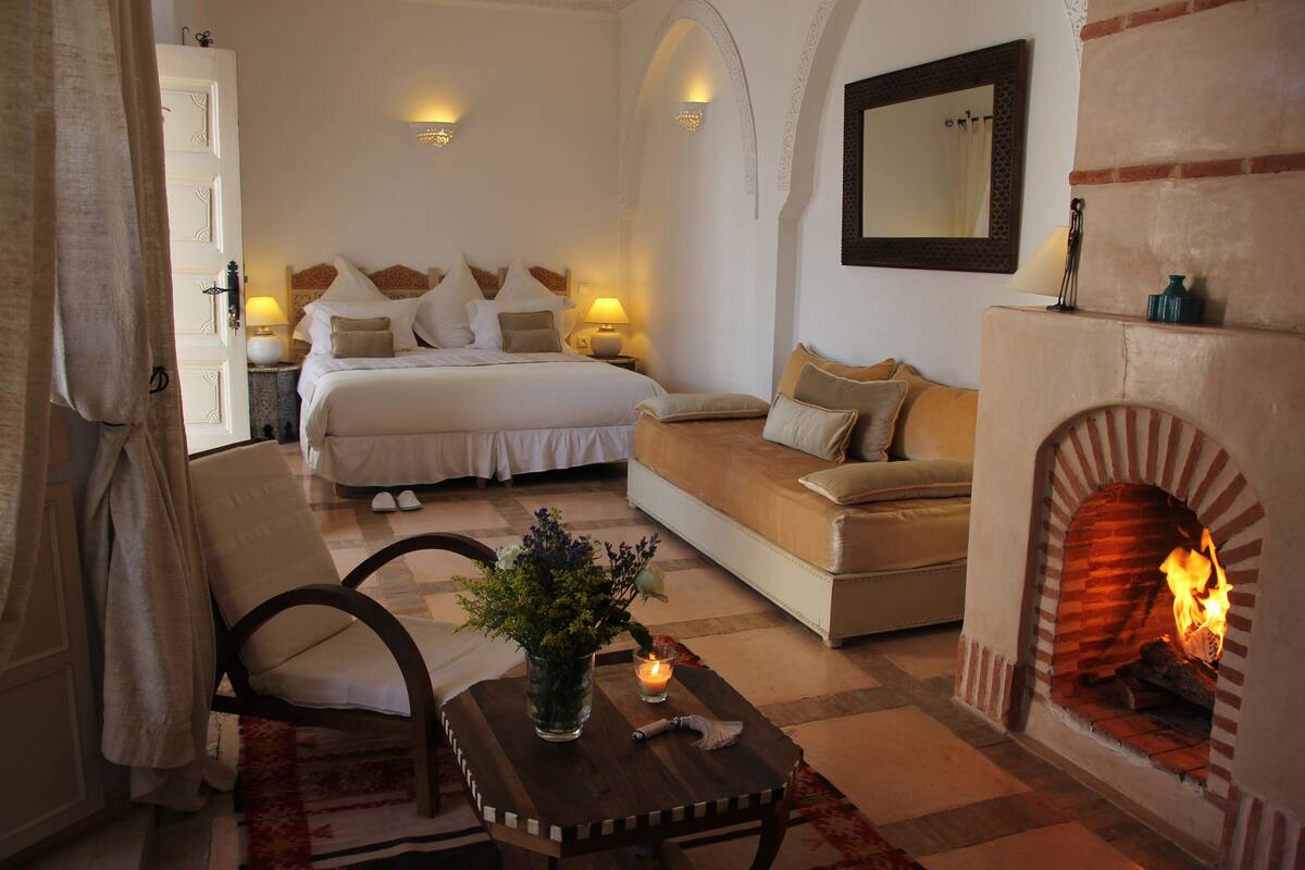 A bedroom with a bed, a sofa, a fireplace, and a nightstand with a lamp.