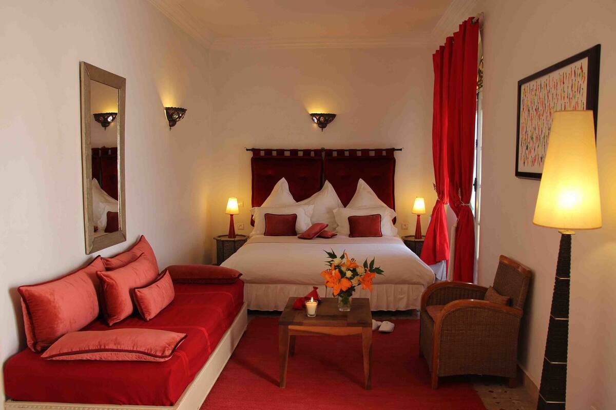 A hotel room with a large bed, red curtains, a red sofa, a small table with a vase of flowers, and a chair.