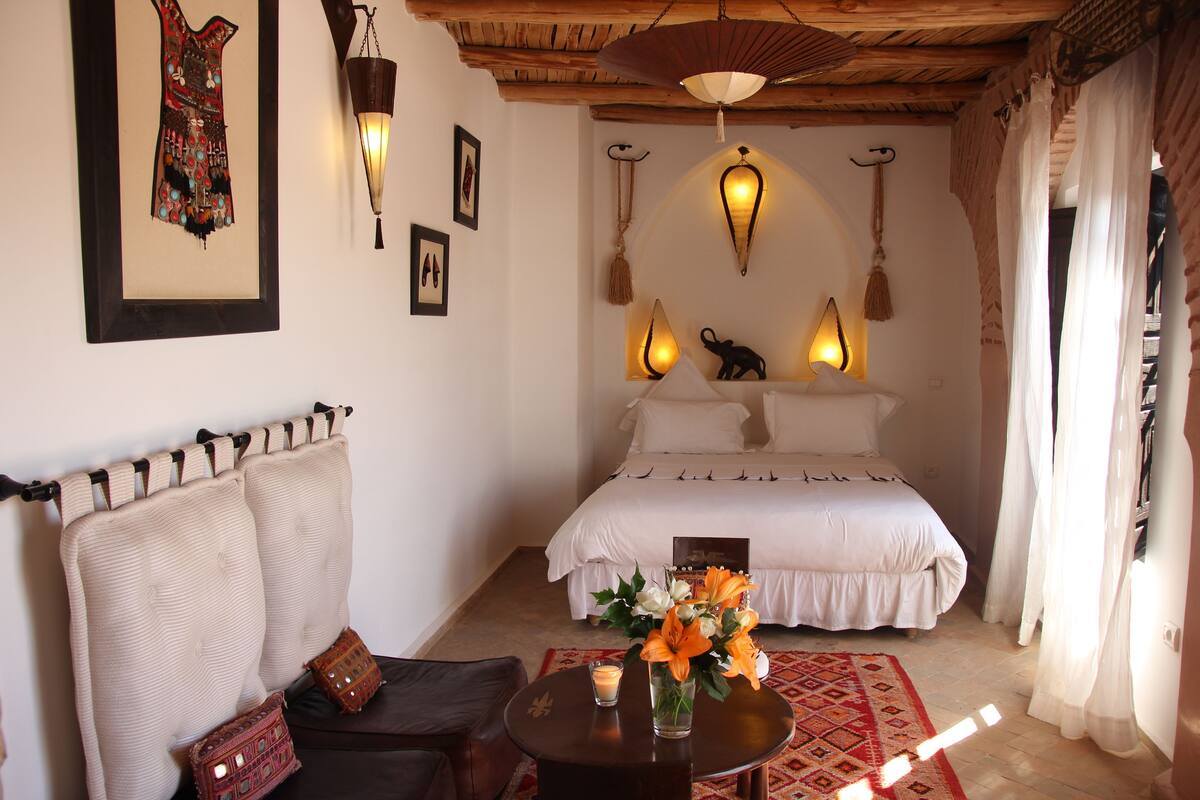 A bedroom with a bed, a nightstand, a chair, a table with a vase of flowers, and wall-mounted lights.
