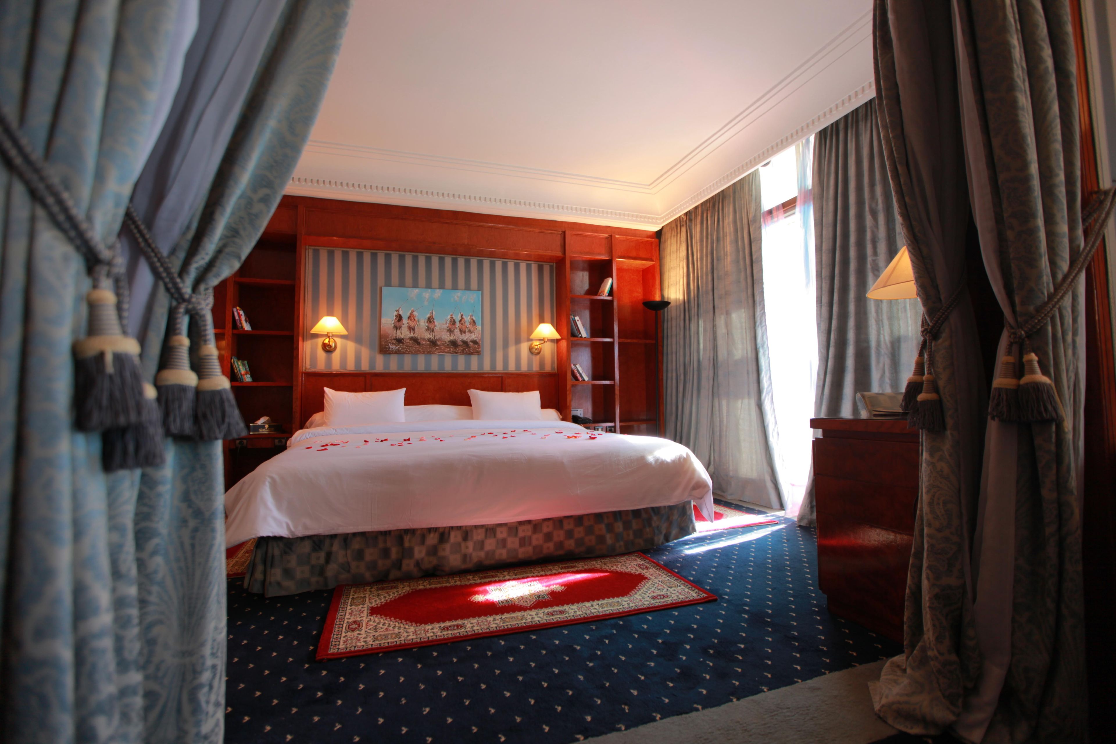 A hotel room with a large bed, a painting on the wall, a bookshelf, and a window with curtains.