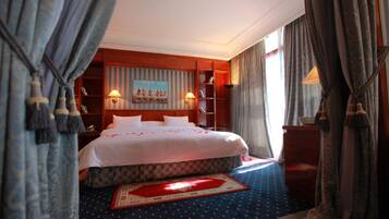 A hotel room with a large bed, a painting on the wall, a bookshelf, and a window with curtains.