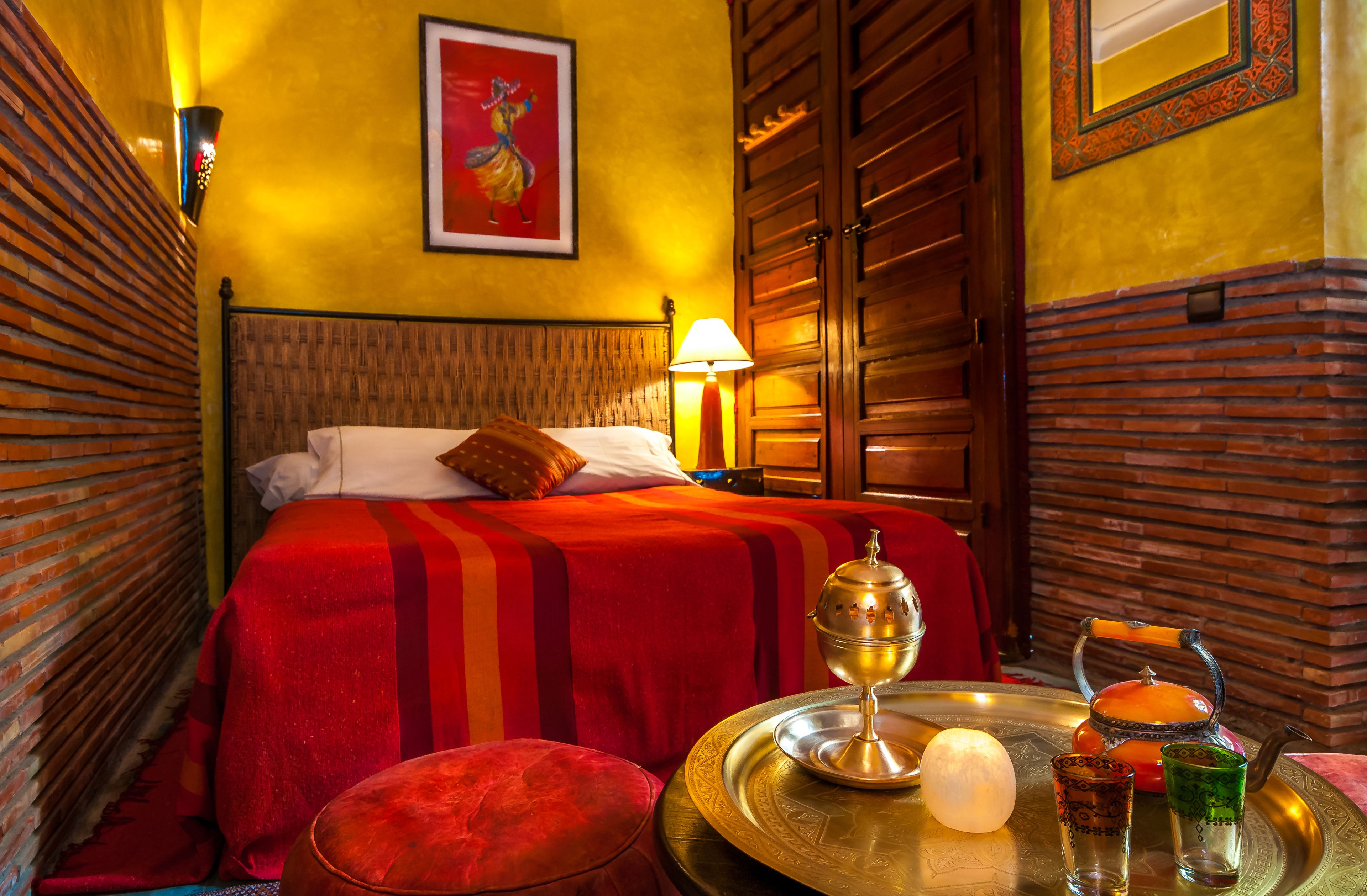A hotel room with a bed, a red and orange striped bedspread, a brass tray with a teapot, glasses, and a candle.