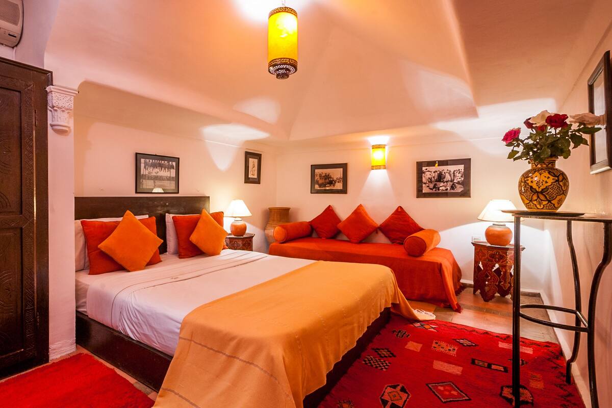 Habitación de hotel con una cama grande, dos mesitas de noche, un sofá, una alfombra roja y una puerta de madera.