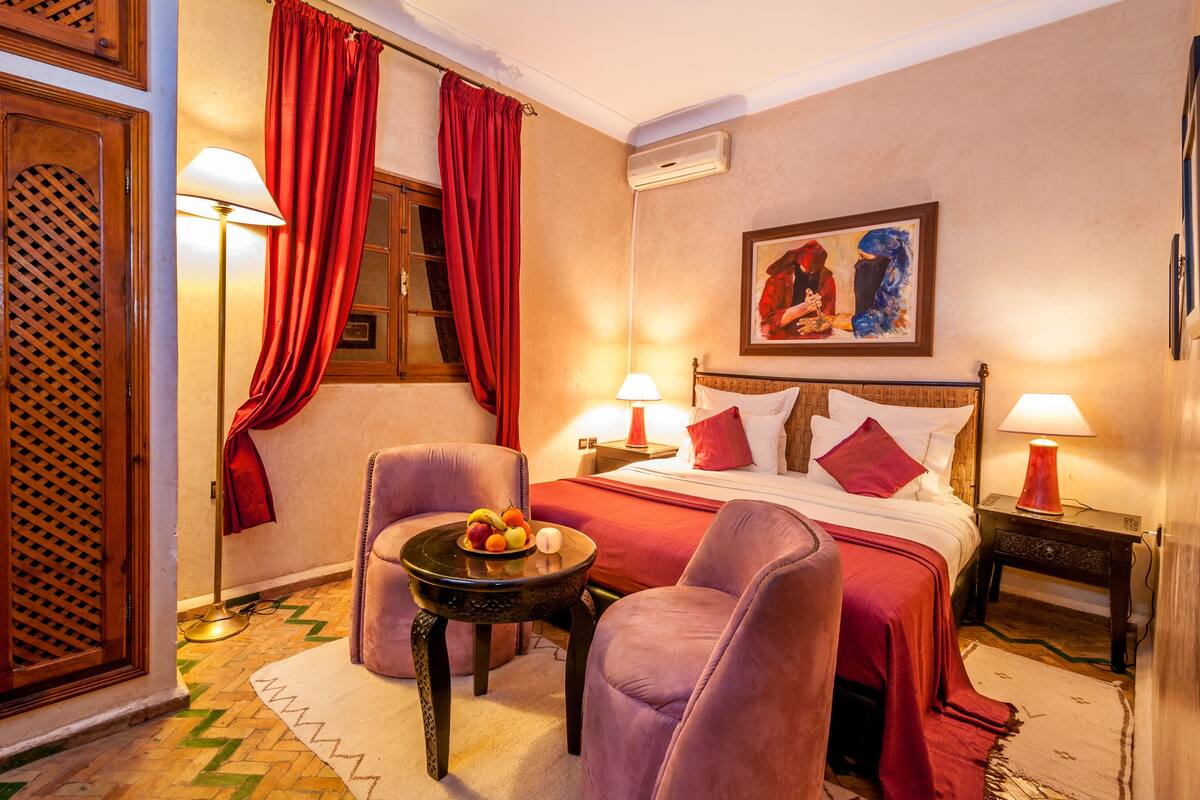 Una habitación de hotel con una cama, dos sillas, una mesita con fruta, una lámpara y un cuadro en la pared.